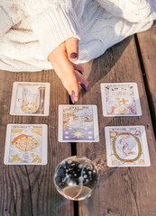 Woman's hand with purple nails points to five Tarot cards spread out on wooden surface next to crystal ball. Vertical