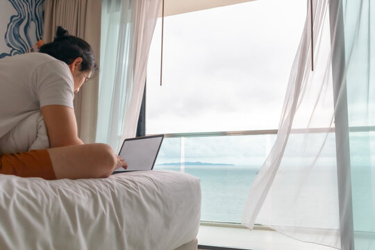 Behind angle of Asian man work on the bed with sea view. Concept of workation.