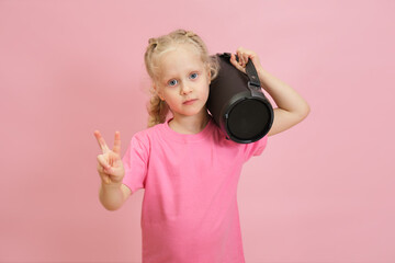 cute preschooler girl with portable wireless speaker on pink background, toddler girl showing peace...