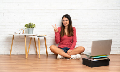 Young woman with a laptop sitting on the floor at indoors smiling and showing victory sign