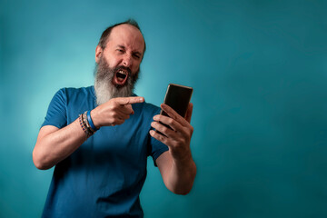 Gray bearded man dressed in blue t-shirt angry, yelling and pointing finger at smartphone