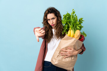 Young woman holding a grocery shopping bag isolated on blue background showing thumb down with...