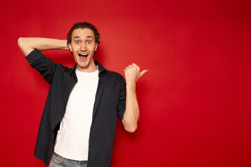 young cheerful surprised confused male student in casual blue shirt points with his index finger to side free work area mock up of copy space isolated on red background studio portrait. promotion