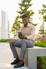 Obraz premium Full length portrait of young handsome African businessman sitting outdoors in city using mobile phone while smiling