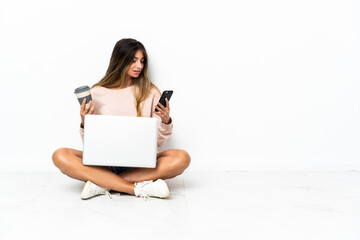 Young woman with a laptop sitting on the floor isolated on white background holding coffee to take away and a mobile