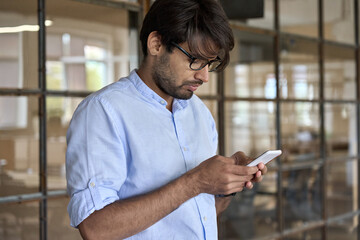 Young Indian business man using cellphone apps standing in office. Arab professional businessman holding mobile phone managing finance banking applications corporate digital technology.