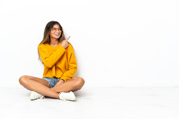 Young woman sitting on the floor isolated on white background pointing finger to the side