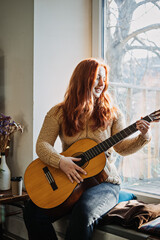 Unaltered candid portrait of young red haired woman in sweater playing acoustic guitar sitting by window at home. Hobbies, indoor home activities for adults in winter, autumn.