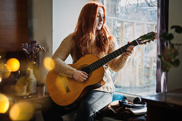 Unaltered candid portrait of young red haired woman in sweater playing acoustic guitar sitting by...