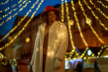 Happy smiling African American woman wearing hat, white faux fur coat, posing with garland at festive Cristmas street fair in European city. Outdoor night portrait. Copy, empty space for text
