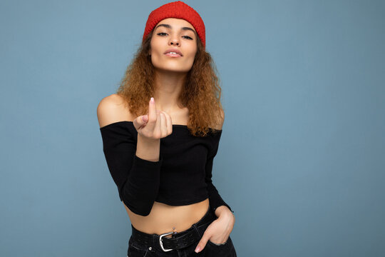 Photo of young positive sexy cute nice brunette woman curly with sincere emotions wearing stylish black crop top and red hat isolated on blue background with copy space and showing beckoning gesture
