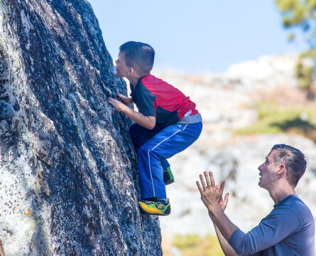 Young boy learning rock climbing techniques from his father who is close by and ready to catch his son if he falls.