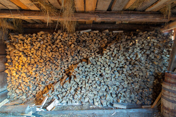 A pile of dry wood lying in an old wooden shed. You can see the walls of the barn, a wooden ceiling with hanging dry hay and an earthen floor. On the right is a barrel and a basket. Soft mixed light.