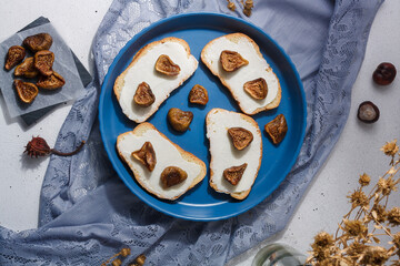 Autumn food composition, cream cheese sandwiches with dried figs. No sugar healthy diet. Blue fabric and dry flowers on white table. Top view