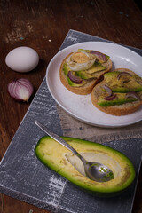 Two sandwiches with avocado, boiled eggs and red onions on white plate, avocado half on wooden background. Vertical shot