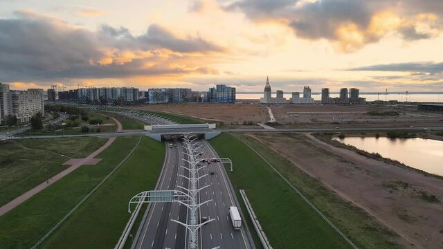 Aerial fly over high speed road in city center. A lots of cars moving fast on highway. Drone is flying up. Sunset, bridge and cityscape on background.