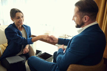 Young business partners smile at each other while shaking hands, sitting in a hotel room after successful negotiations. Young business woman at a meeting with a handsome male business partner