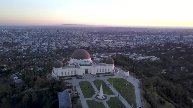 Aerial orbit of the Griffith Observatory and the Los Angeles skyscraper skyline in the background in the evening.