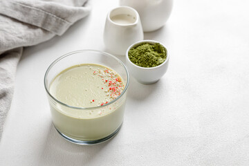 Matcha panna cotta dessert in a glass on a white background and a napkin next to it. Milk jug and matcha in the background.
