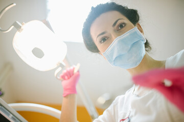 Female dentist in dental office examining patient teeth, camera angle from patients perspective. 