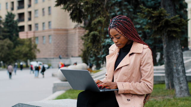Serious african american girl blogger using laptop, sitting outdoor, young busy focused businesswoman looking at screen, intern typing on keyboard, writing report, student working on research project