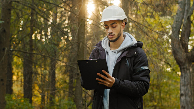 Young experienced Indian man forestry engineer in hardhat with tablet taking measures for reforestation of woodlands. Working and Supervising in park, watching of wildlife sanctuary checking trees