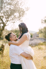 Groom holds bride in his arms against the background of trees on the lawn