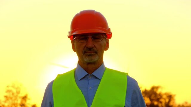 Technician in uniform on windmill substationat sunset. Worker in orange hardhat walks at construction site against rotating turbine closeup