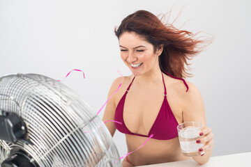 Naklejka premium Red-haired smiling woman in a bikini drinks a cold drink and enjoys the blowing wind from an electric fan on a white background. Climate control on a hot summer day