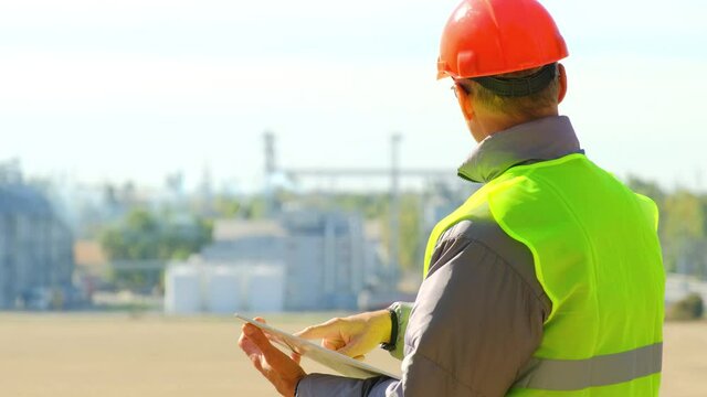 Grain elevator farm worker. Concentrated man builder looks at drawings on tablet inspecting new food factory with large silo tanks in field