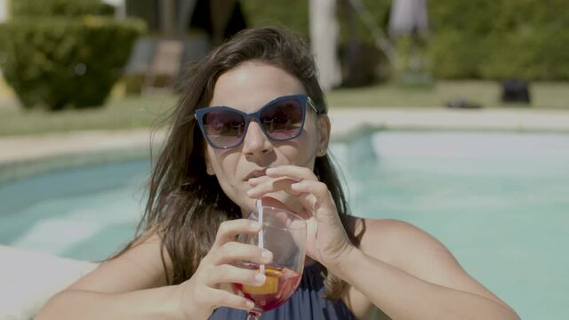 Front view of happy woman drinking cocktail in swimming pool. Medium shot of attractive Caucasian girl in swimsuit and sunglasses relaxing on summer day, looking at camera and smiling. Party concept