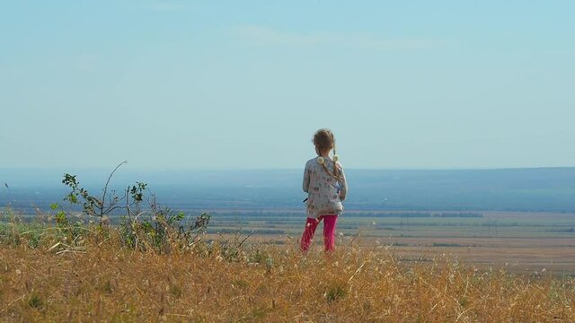 A girl with pigtails stands on top of a hill and looks into the distance and strong wind shakes the grass, girls hair and her dress.