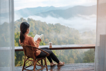 young woman reading book near window and looking mountain view at countryside homestay in the...