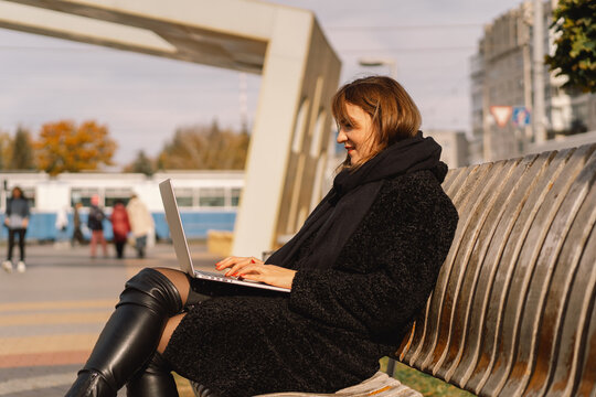 A woman sits on a bench in the city, uses a computer and talks on a video call. Freelance woman. People and technology
