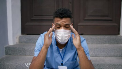 Frustrated male doctor sitting in front of the building with face mask and looking at camera.