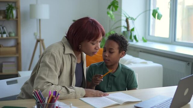 Online home school for junior schoolchildren. Little black boy hugs mother and presses key to answer videocall via laptop at desk in living room spbd