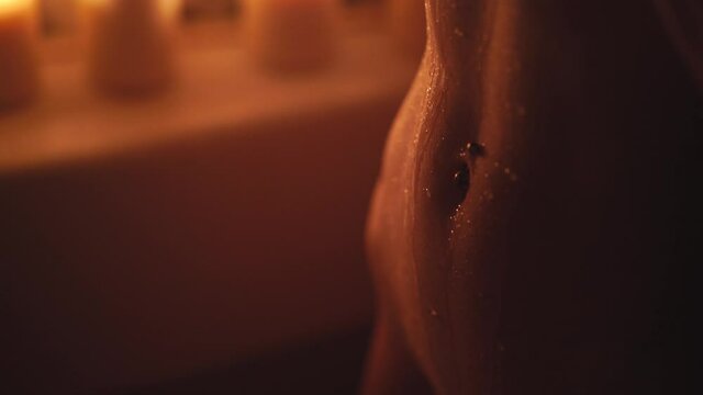 A young girl with freckles in a dark bathroom with candles pours water on her belly. Navel piercing