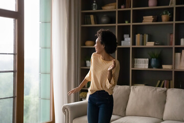 Joyful excited African American girl having fun, enjoying fitness activity, exercises at home, dancing, laughing, singing along to music on radio. Young homeowner woman celebrating good event