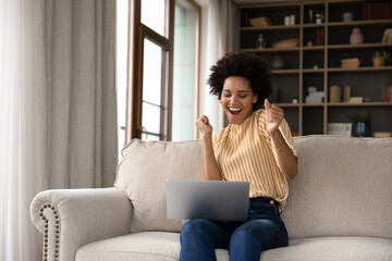 Excited cheerful young Black woman using laptop computer on sofa at home, getting good news,...