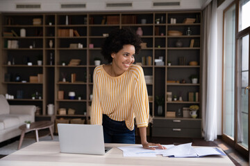 Happy Black millennial remote worker satisfied with job result looking at window away, smiling, thinking over good startup project future vision, dreaming on career success at workplace with laptop
