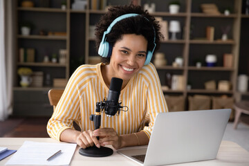 Happy cheerful young Black woman in headphones and professional microphone recording audio podcast...