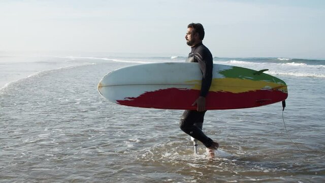 Long shot of steady surfer with bionic leg going to shore. Side view of happy sportsman with surfboard in hands finishing surf workout during vacation on seaside resort. Sport, challenge concept