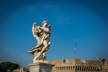 Fototapeta premium Angel with the Superscription on Ponte Sant'Angelo (Bridge of Hadrian) in front of Castel Sant'Angelo (Castle of the Holy Angel) in Parco Adriano, Rome, Italy