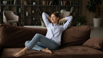 Serene young 30s woman relaxing on couch enjoy carefree weekend alone at modern cozy home. Concept of happy homeowner spend day off inside comfort apartment with air conditioner and cosy furniture