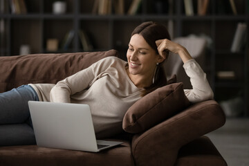 Relaxed young smiling woman rest on couch with laptop, watch movie on-line, make order, purchasing goods on internet for home, spend weekend alone at apartment, use modern tech, enjoy leisure concept