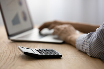 Close up focus on calculator, on background female hands on laptop keyboard, graphs charts on pc monitor. Banking, stats application services usage, investments, management of budget, finances concept