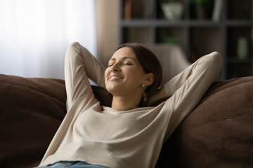 Close up of serene peaceful smiling 30s woman lean on sofa with hands behind head closing eyes...