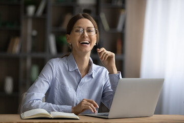 Fototapeta premium Happy attractive 35s businesswoman in glasses sit at workplace desk in front of computer laughing feels overjoyed got great online news. Portrait of successful independent female entrepreneur concept