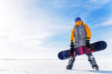 Young man holds a snowboard in his hands against the background of a winter landscape. Male in ski goggles and overalls with his board on white snow on a sunny frosty day go in for sports.