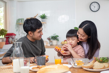 Asian family breakfast at home. Parents and children enjoy eating together, talking with laughter and good atmosphere. Father plays with son playfully at kitchen table.